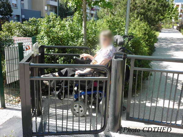 Une personne handicapée, se déplaçant en fauteuil roulant électrique, est rentrée dans une chicane mobile placée au bout de l’allée d’un parc. Cette chicane est formée d’un grand « U » métallique qui tourne autour d’un axe vertical. Le fauteuil a des cale-pieds, mais les pieds dépassent, et ne sont pas protégés. La personne ne peut pas faire pivoter la chicane sans aide. Le fauteuil roulant reste bloqué dans la chicane et la personne handicapée semble être prisonnière….